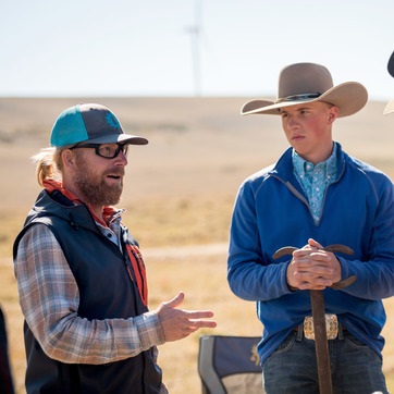 Two men stand outdoors talking; one wears sunglasses and a cap, the other a cowboy hat and blue jacket, holding a wooden stick. A wind turbine is visible in the background.
