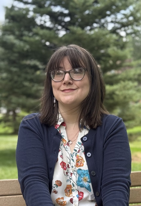 Ashley Laughlin sitting on a bench in front of trees wearing a blue sweater and floral patterned shirt.