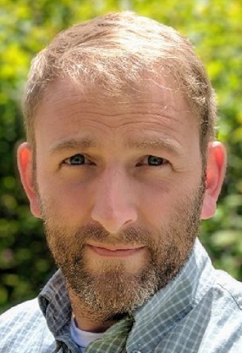 Chris Rothfuss wearing a blue and white striped shirt in front of green foilage.