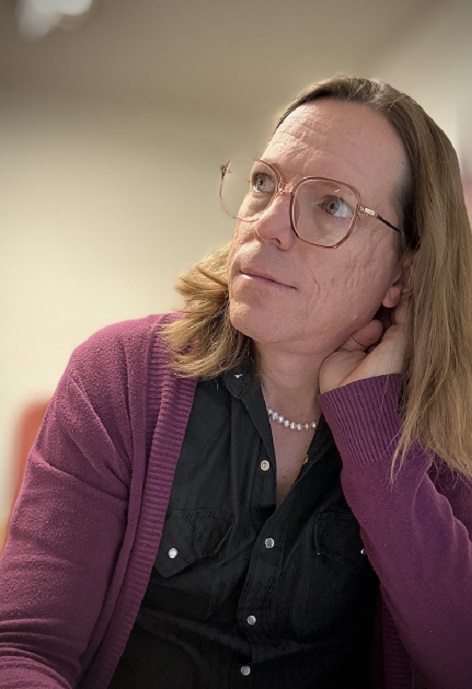 Ella Daniel Gray wearing a black shirt and pink sweater in front of a blurred white background.