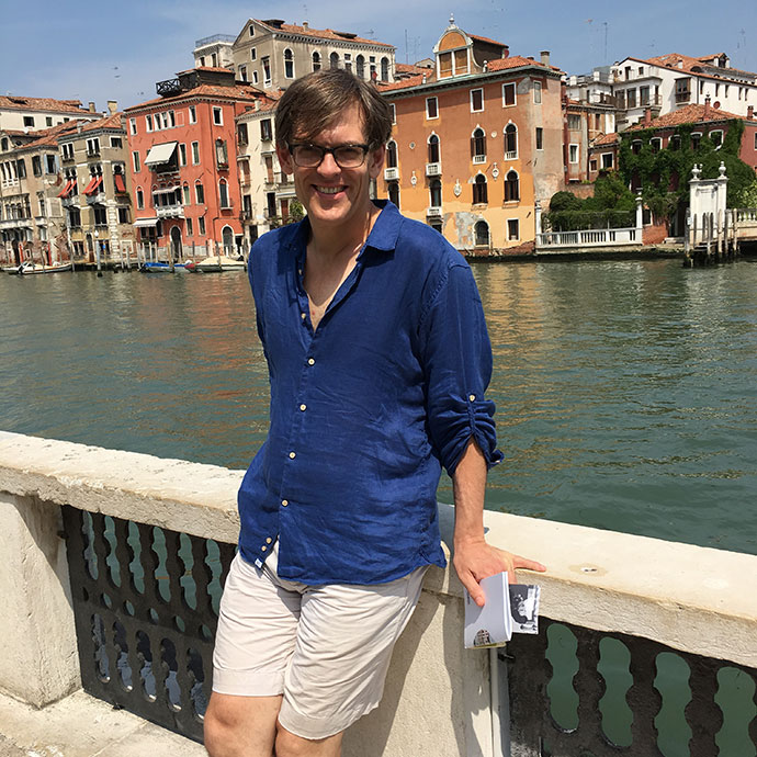 man leaning against a low wall with water and buildings behind him