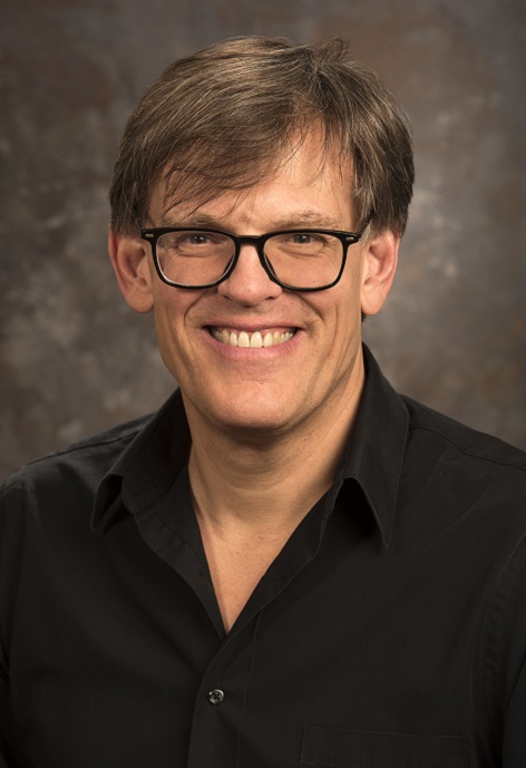 Peter Parolin wearing a black shirt and glasses in front of a grey background.