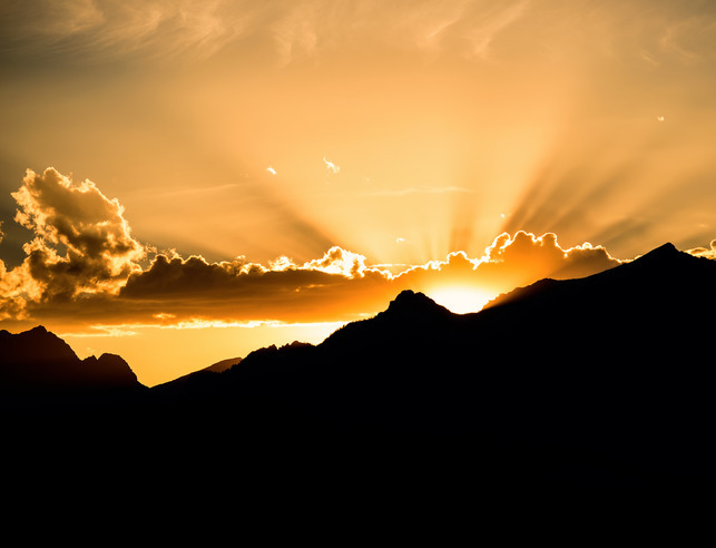 A sun rising over a mountain landscape in dark shadow.