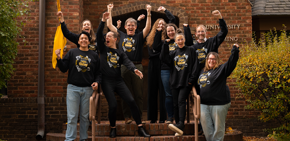 Honors faculty and staff cheer together in front of the Guthrie House wearing black and yellow sweatshirts that say "You Belong."