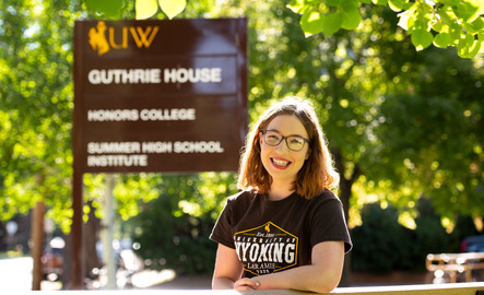 A honors student broadly smiles in front of the Honors House sign on the University of Wyoming campus.