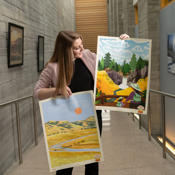 Student holding two landscape paintings on paper in a grey hallway.