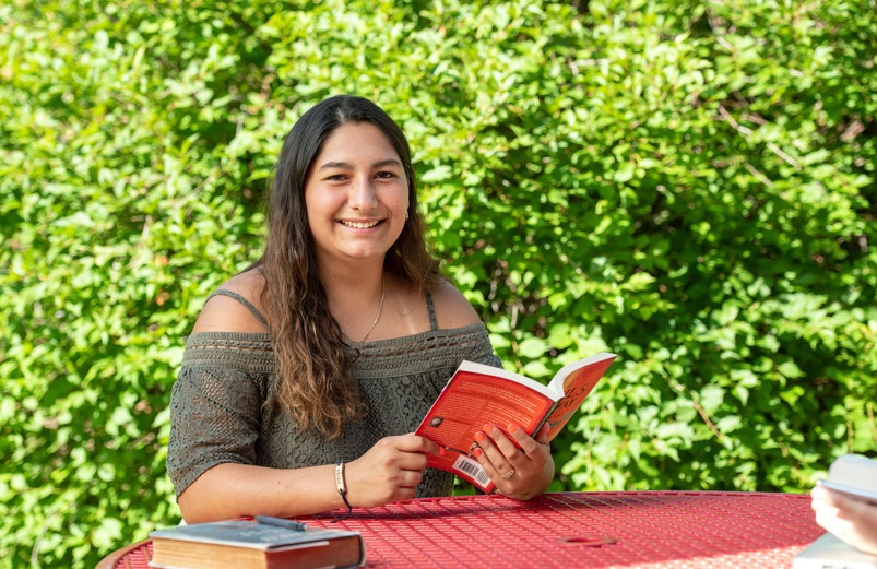 A person sits at a red table outdoors, reading a book, surrounded by lush green foliage.