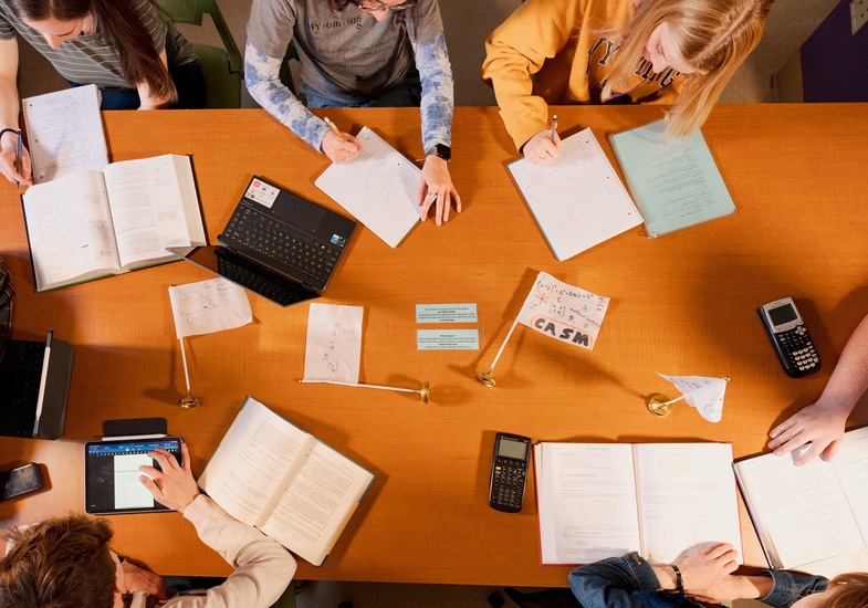 Aerial view of a study session with students writing notes, textbooks, laptops, and calculators on a wooden table.