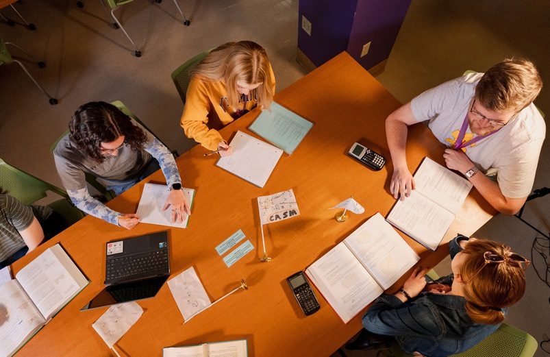 Five students sit around a wooden table with open notebooks, laptops, and calculators, focused on studying. The atmosphere is collaborative and studious.