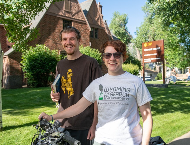 Two students stand beside a bicycle on a grassy area in front of a brick building, showcasing a campus atmosphere at the University of Wyoming.