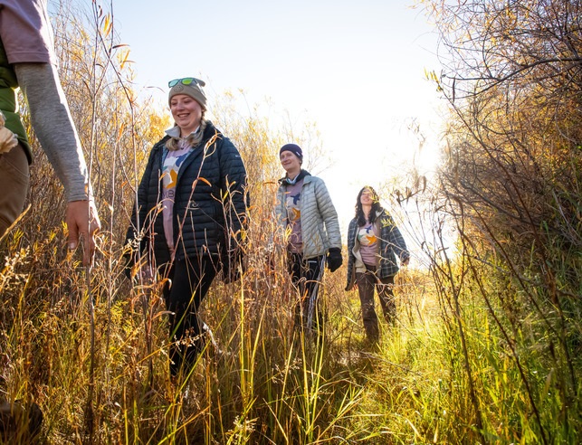 Four people smiling as they walk through a sunlit, grassy trail surrounded by autumn foliage. Their expressions convey joy and a sense of adventure.