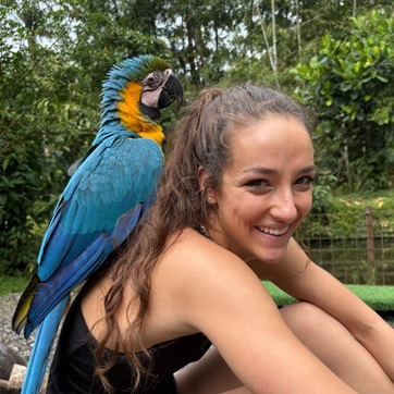 Student sitting surrounded by green fiolage with a blue and yellow bird sitting on her shoulder.