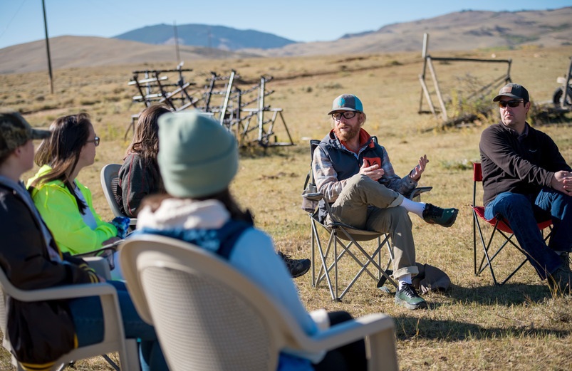 Faculty member talking to a group of students outdoors sitting in camping chairs