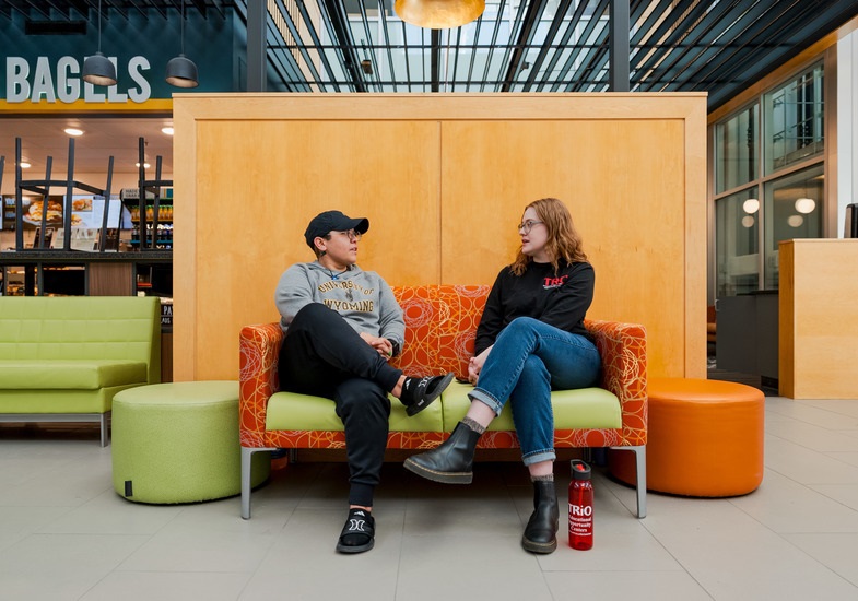 Two people sit on a colorful sofa in a modern space, surrounded by furniture and a café setting, engaged in conversation.