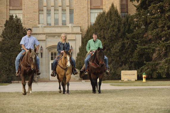 3 people on horses in front of the Ag building. Perpetuating the myth that Wyoming rides horses to work.