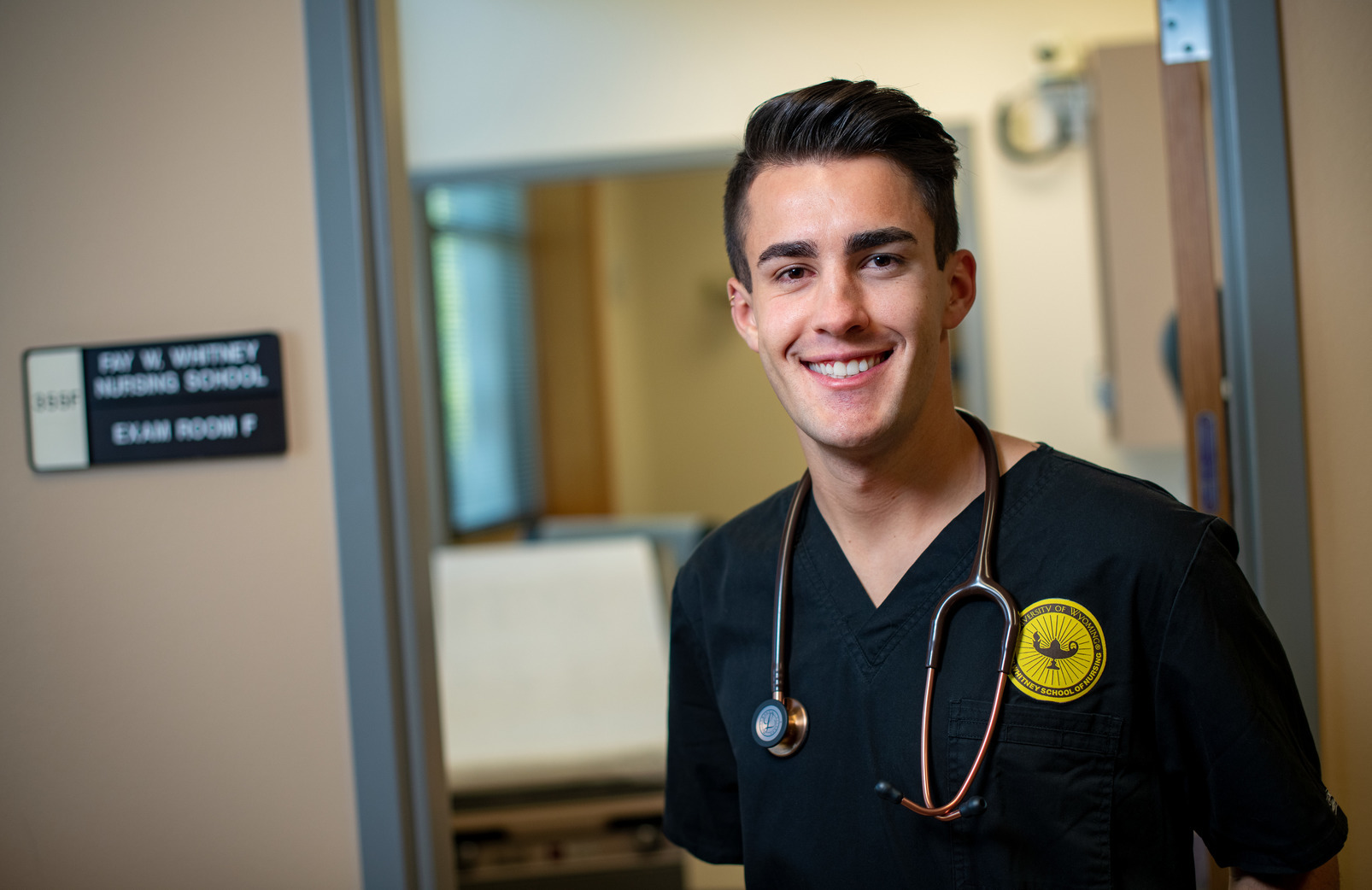 A nursing student in scrubs standing in front of an exam room.
