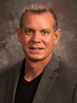 A man with short brown hair wearing a gray suit jacket and a black t-shirt.