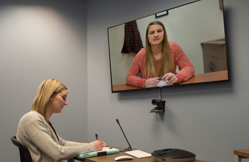 A person sitting at a desk taking notes, while a TV above them shows another person talking.