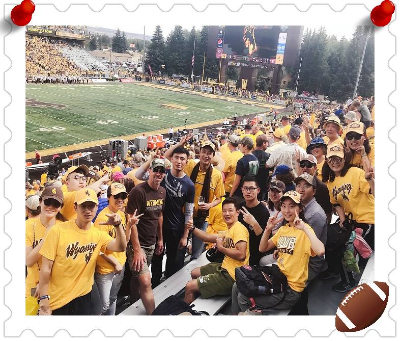 A large group wearing yellow shirts with "Wyoming" on the front smile and wave at the camera, there is a football field in the background.