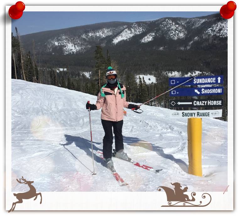 A woman in skiing gear standing next to a sign, there is snow on the ground and pine tree covered mountains in the background.