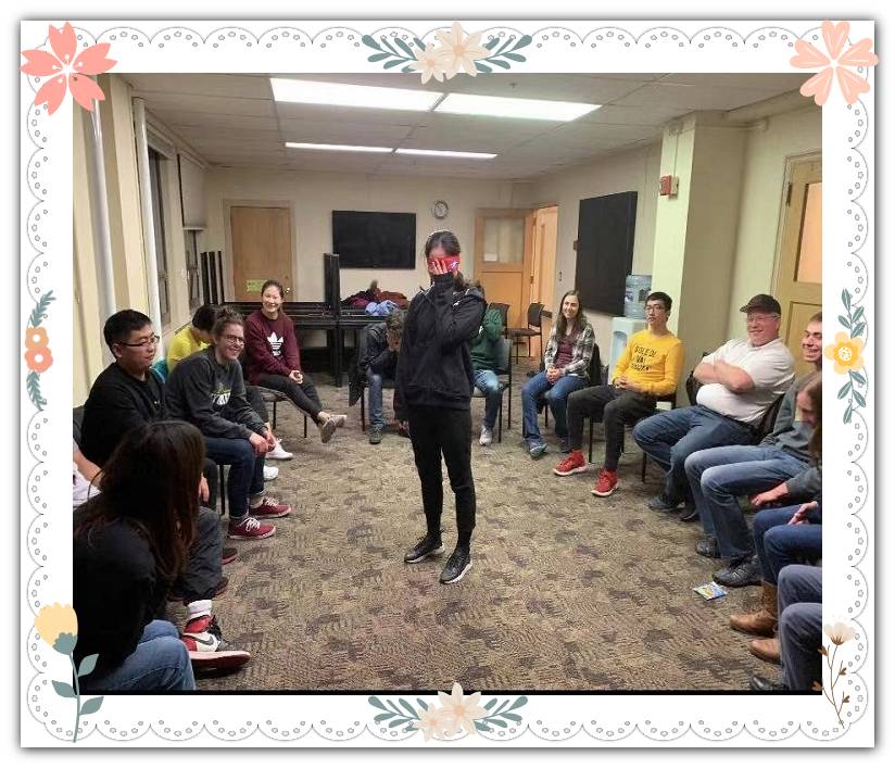 A person stands blindfolded in the center of a circle of chairs that have people sitting in them.