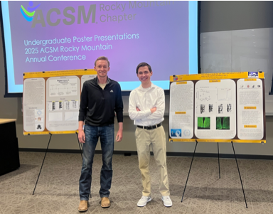 Two people standing on a stage with research presentation posters behind them.