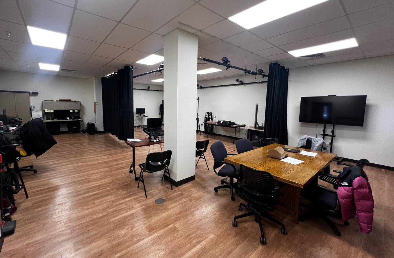 A room with white walls and laminate wood flooring. There are various tables, chairs, and motion tracking equipment.