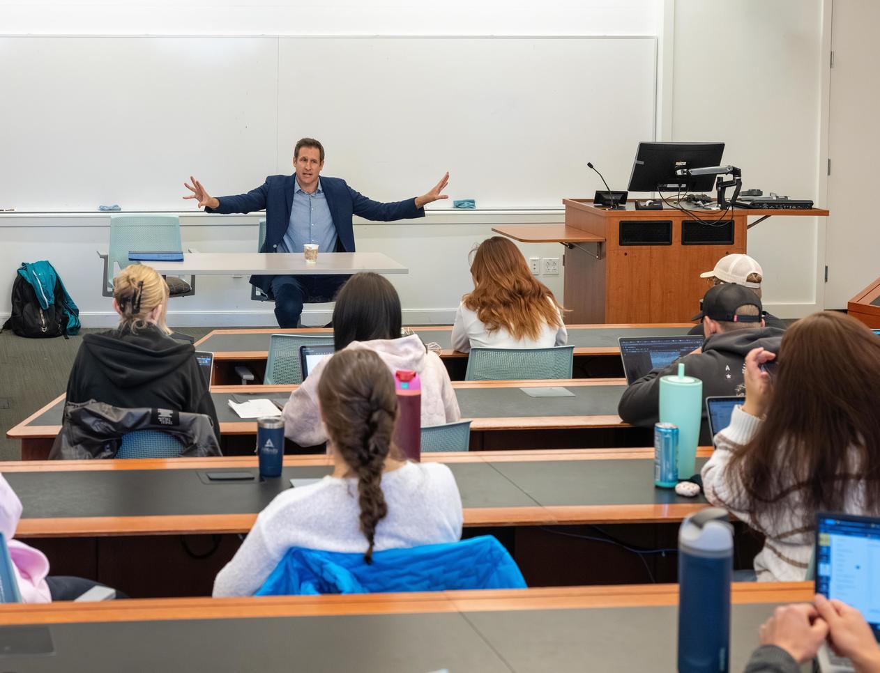 a professor sits at the front of a classroom while students are taking notes