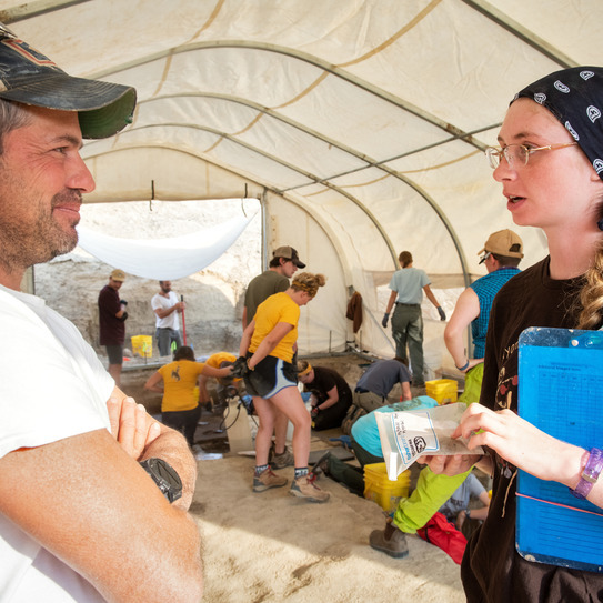 Anthropology research professor and student meet inside tent with other researchers in the background.