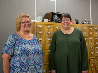 Dee Salo and Debbie McCarthy in the RDM office in front of and old card catalog holder