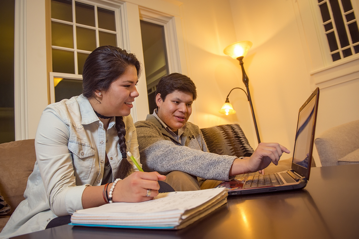 A picture showing a boy pointing at a laptop screen and a girl writing in a notebook with a penicl. 