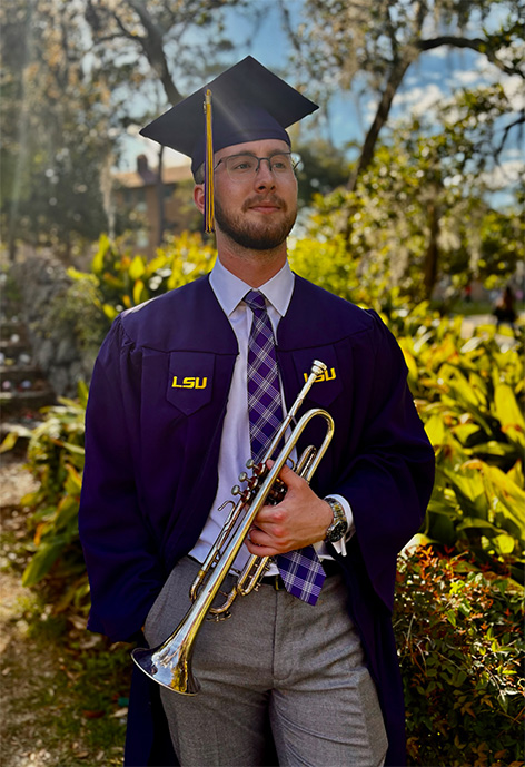 Man in a lavender shirt and purple tie wearing a purple graduation cap and gown holding a trumpet in front of plants