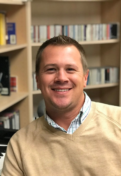 Picture of man smiling wearing tan sweater in front of bookshelf
