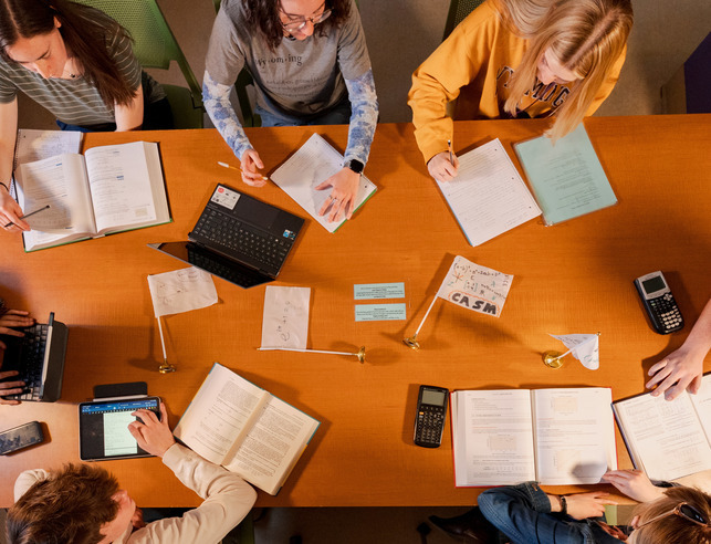 An aerial view of students sitting with open books and laptops at a desk with a small flag that reads "CASM" at the tutoring center 