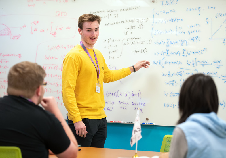 A tutor smiles while pointing at a whiteboard in front of two students