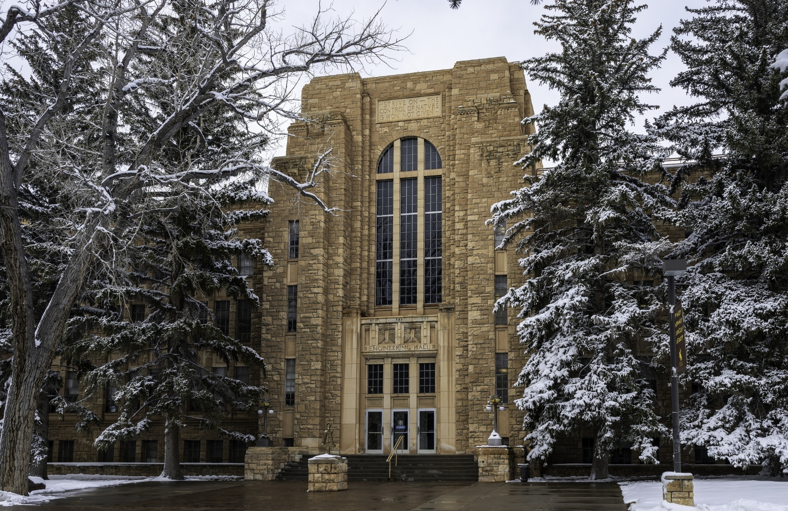Photo of the front of the engineering Hall after a spring snow. The sky is grey and the trees and lawn are coated in snow but the sidwalk has melted.