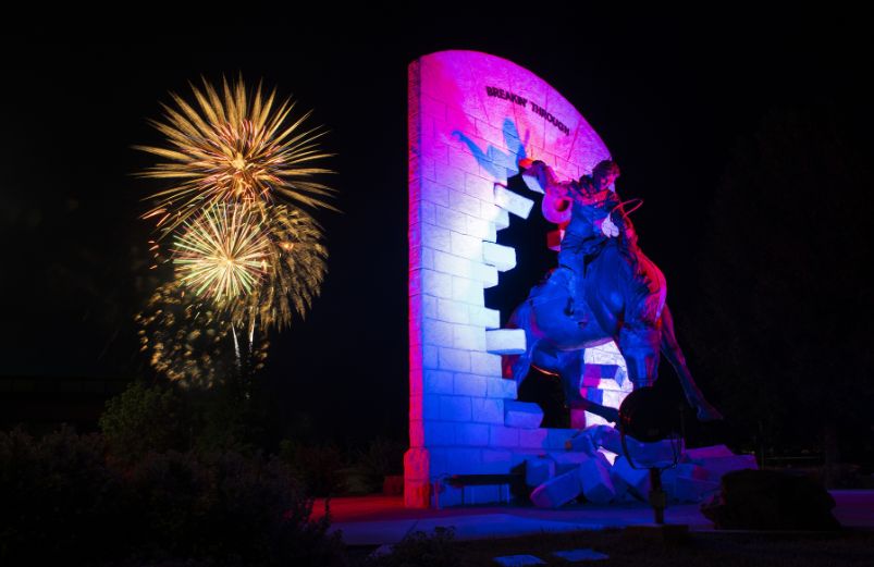 photo of statue "breaking through" lit with blue and red lights with fireworks in the background