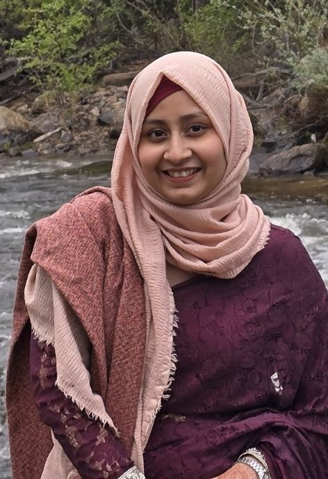 a candid photo of sadia afrose in a burgundy lace dress and pink head scarf, taken outdoors with a stream and trees in the background