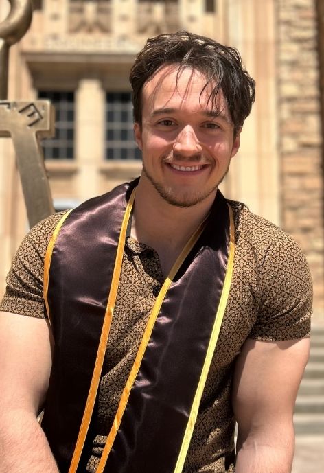 candid photo of austin garcia, wearing a brown and gold patterned polo shirt and the sash from his B.S. Graduation, taken outdoors in front of the UW engineering hall next to the Tau Beta Pi statue.