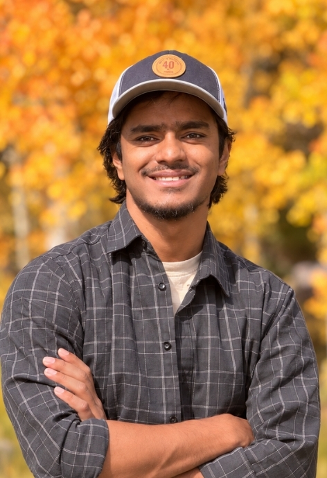 candid photo of Jahid Hasan wearing a plaid button up over a t-shirt and a ball-cap, taken outdoors in with fall trees in the background.