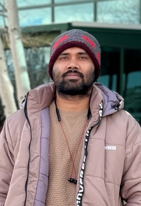 candid photo of mahruf hossain taken outdoors, he is wearing a light pink/beige puffer jacket and sweater and a grey and red knit cap.