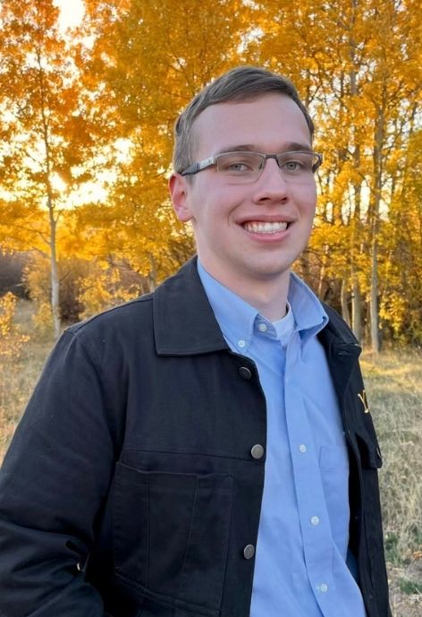candid photo of grant litynski wearing a black denim jacket over a blue button up shirt, taken outdoors with aspen trees with gold foliage in the background.