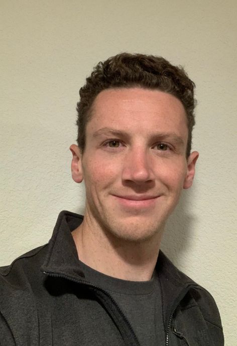 candid photo of aiden mcdonald wearing a grey zip up jacket over a grey shirt in front of a beige background.