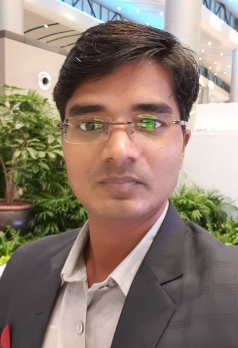 candid photo of ram mohato wearing a grey suit jacket over a white shirt, taken outdoors in front of a white building and shrubbery.
