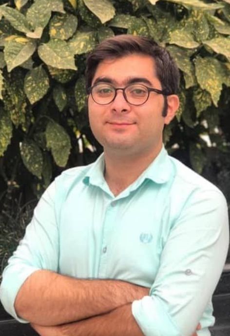 candid photo of mostafa ojaghloo wearing a light blue button up shirt, taken outdoors in front of shrubbery.