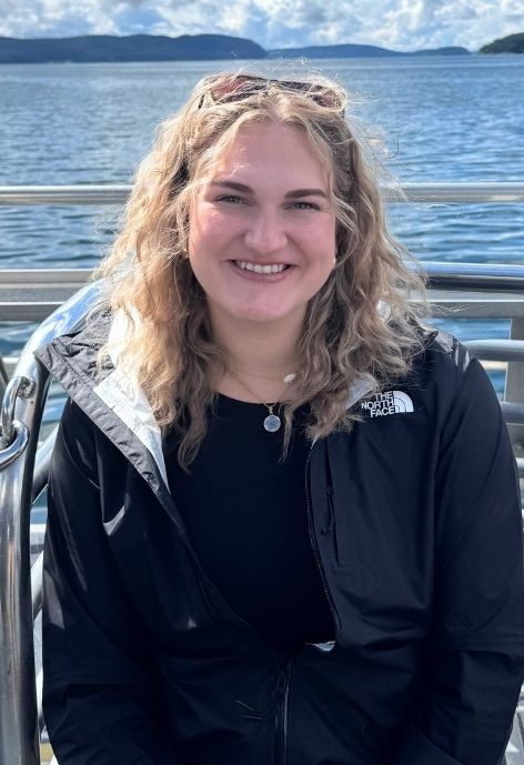 candid photo of erin poyer wearing a black north face brand rain jacket over a black shirt, taken outdoors with a lake and mountains in the background
