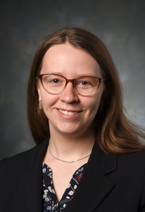 professional headshot of rebekah saxena. she is wearing a black blazer over a dark printed blouse, the background is dark grey.