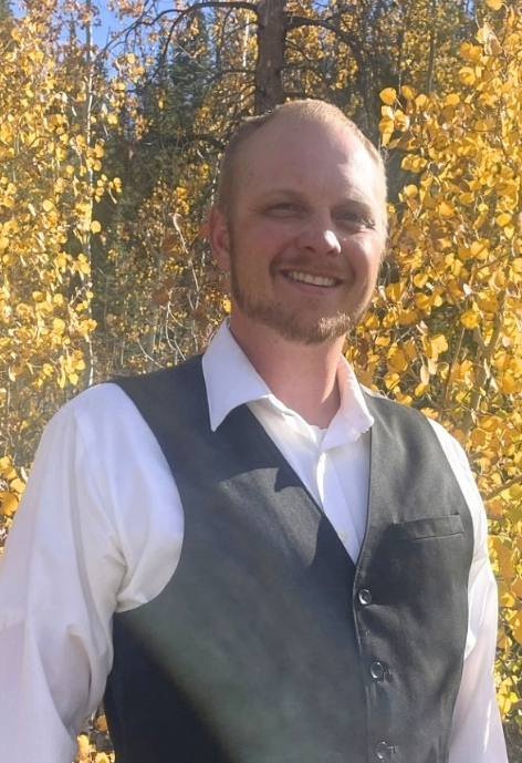 candid photo of keith schnieder wearing a black vest over a white dress shirt and navy patterned tie in front of a colorful background.