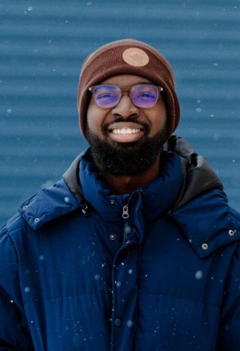 candid photo of charles yeboa wearing a dark blue puffer jacket and brown knit cap, taken outdoors during light snowfall in front of a medium blue corrugated metal background.