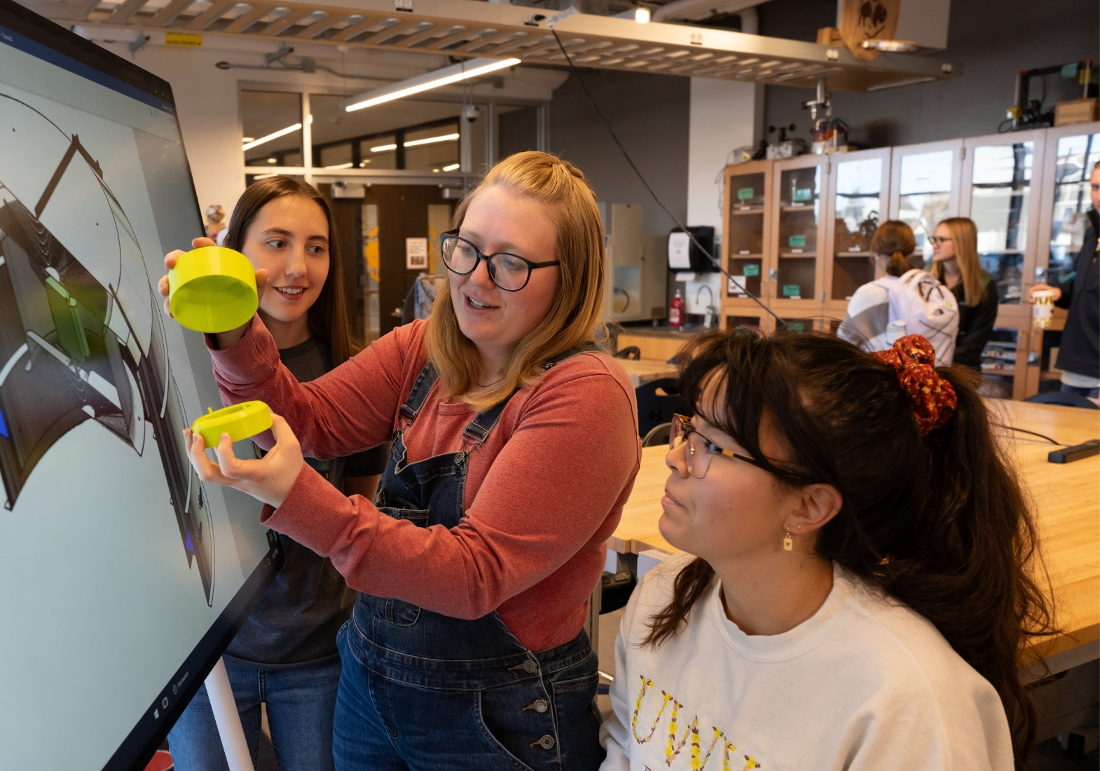Three M.E. senior design students gather around a large monitor displaying an image of the a model design while the center student assembles the components of the prototype.